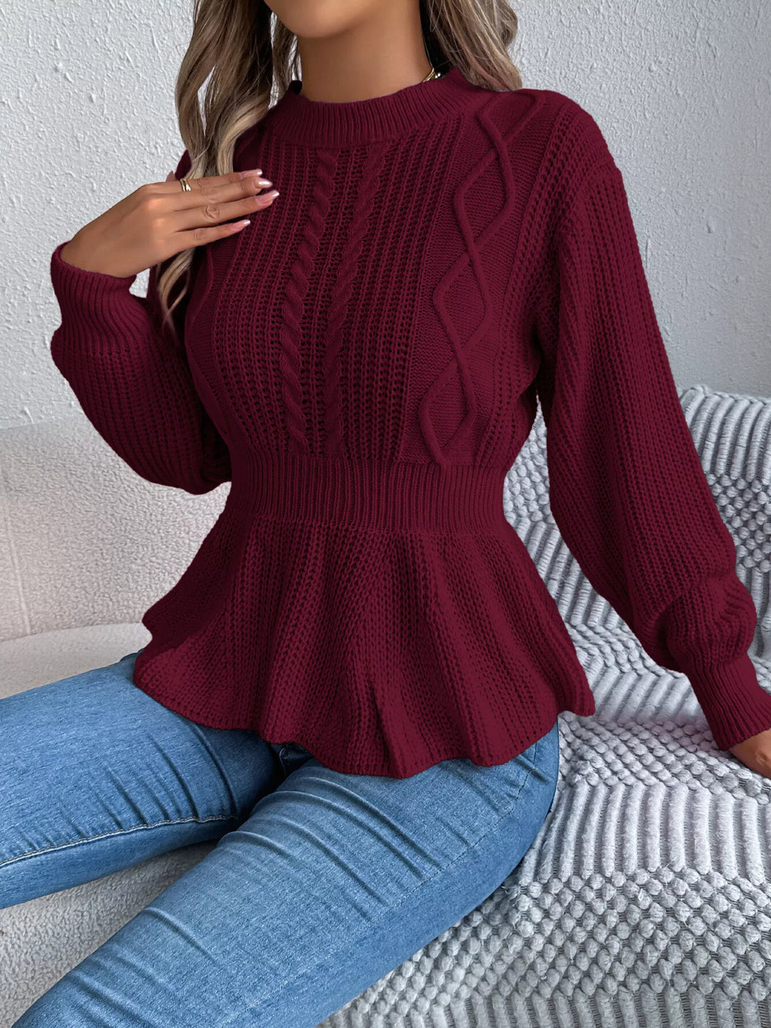 Burgundy cable knit sweater with peplum hem, model seated in jeans