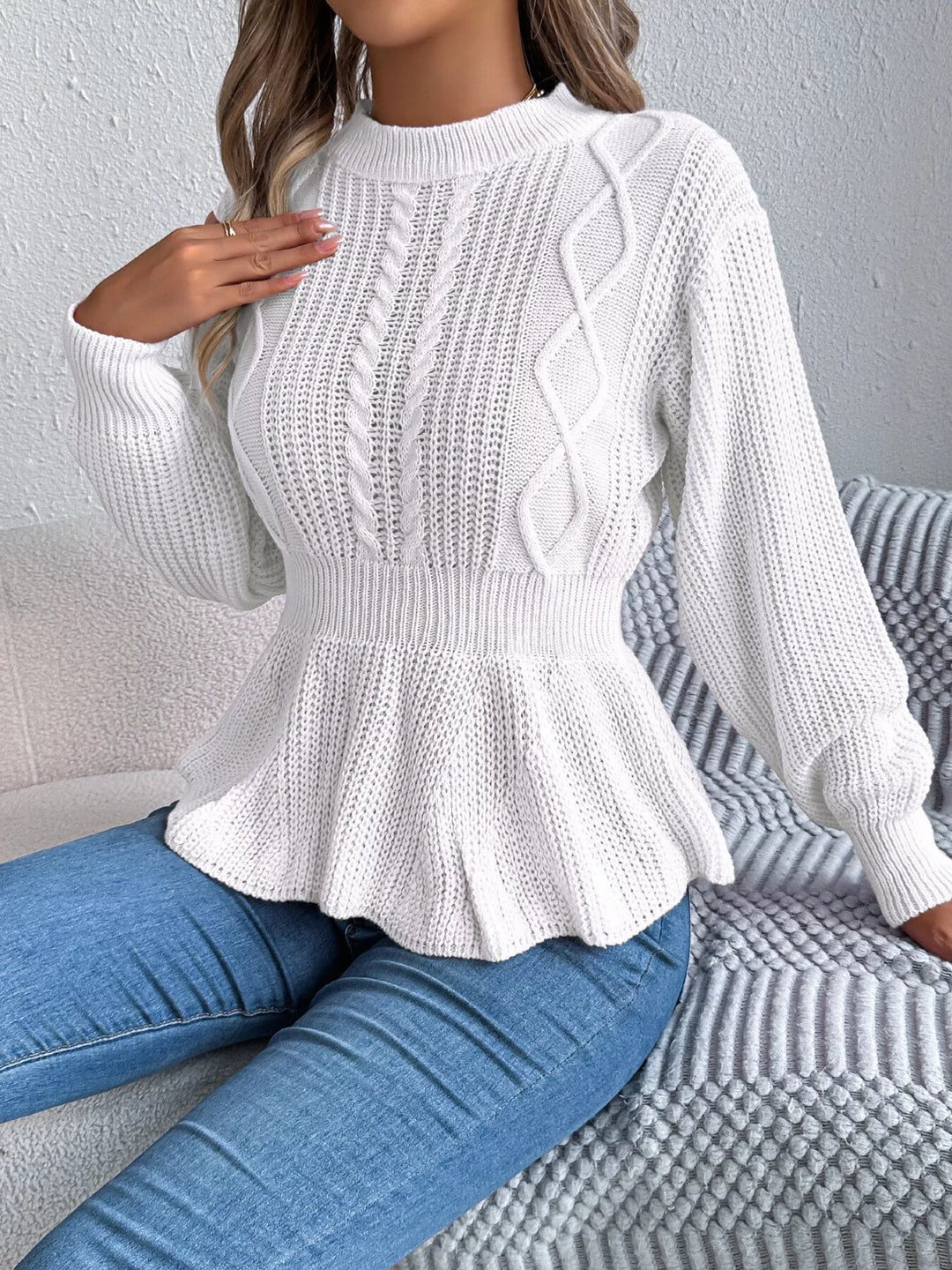 White cable knit sweater with peplum hem, model seated on a gray sofa