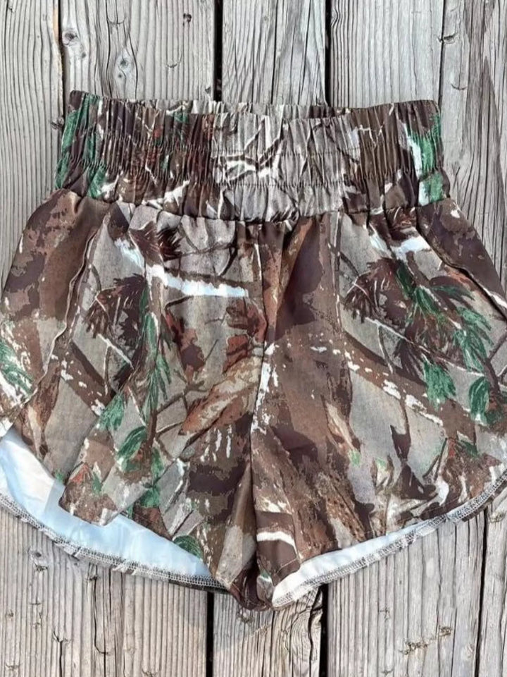 Forest camo shorts with tulip hem laid flat on a weathered wood surface