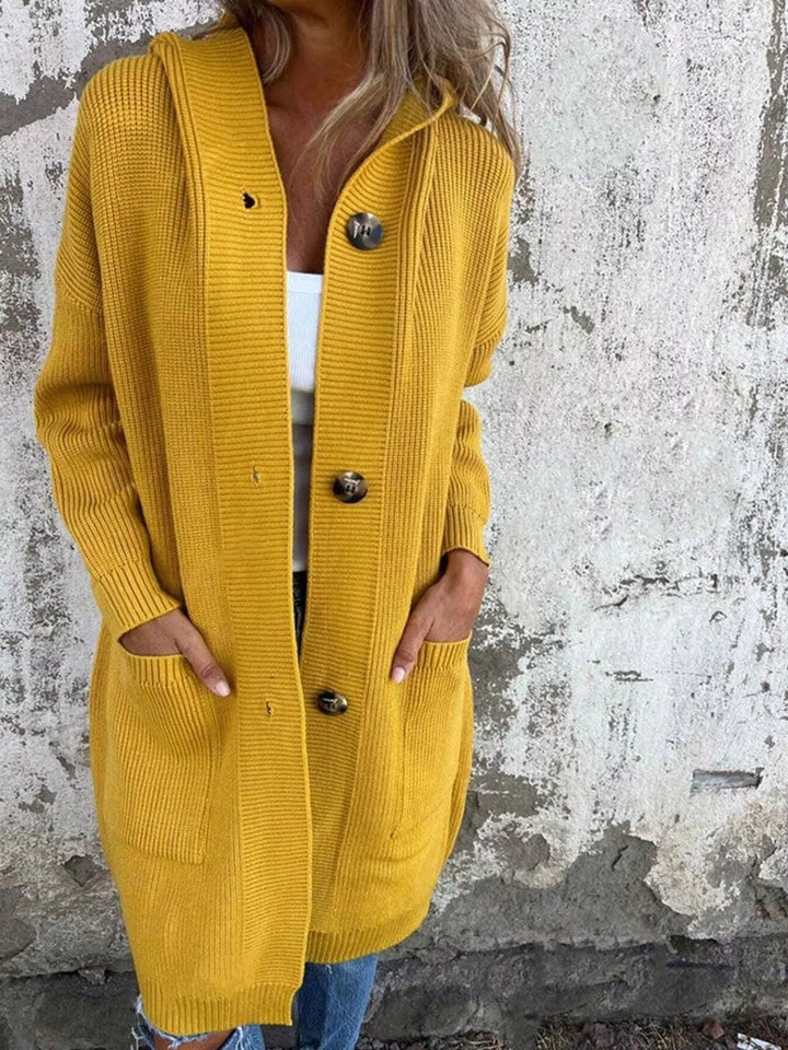 Mustard long hooded cardigan worn with hands in pockets against a concrete wall