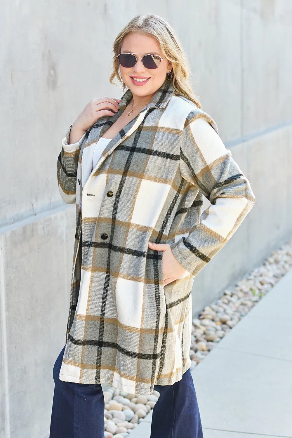 Olive plaid coat with open front and hands in pockets against a concrete wall