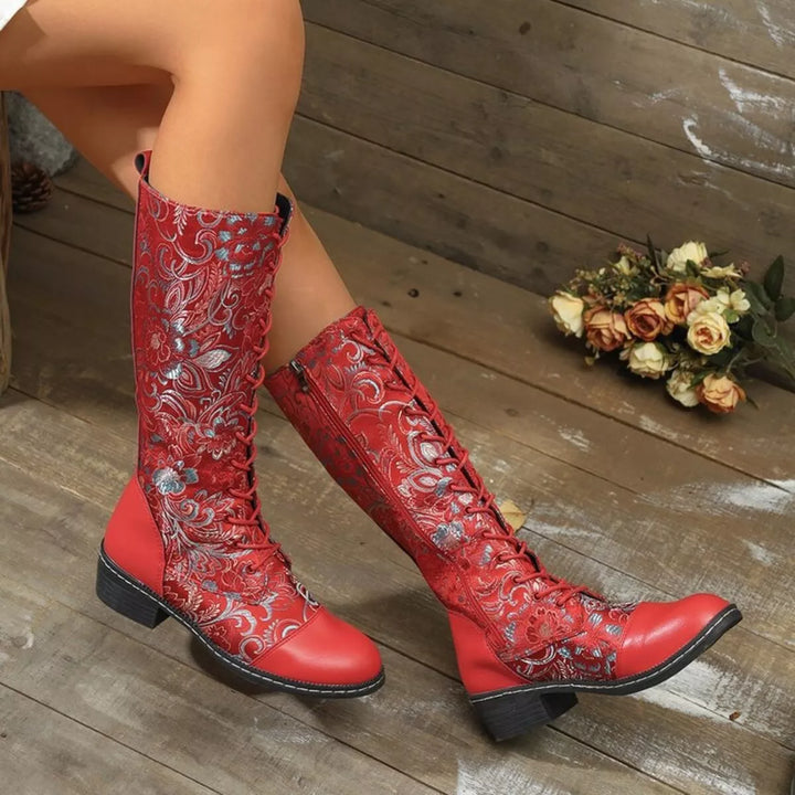 Red mid calf lace up boots worn with legs crossed, floral embroidery visible