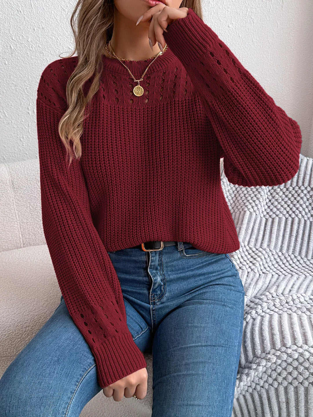 Burgundy knit pullover worn seated on a sofa with a belted pair of jeans
