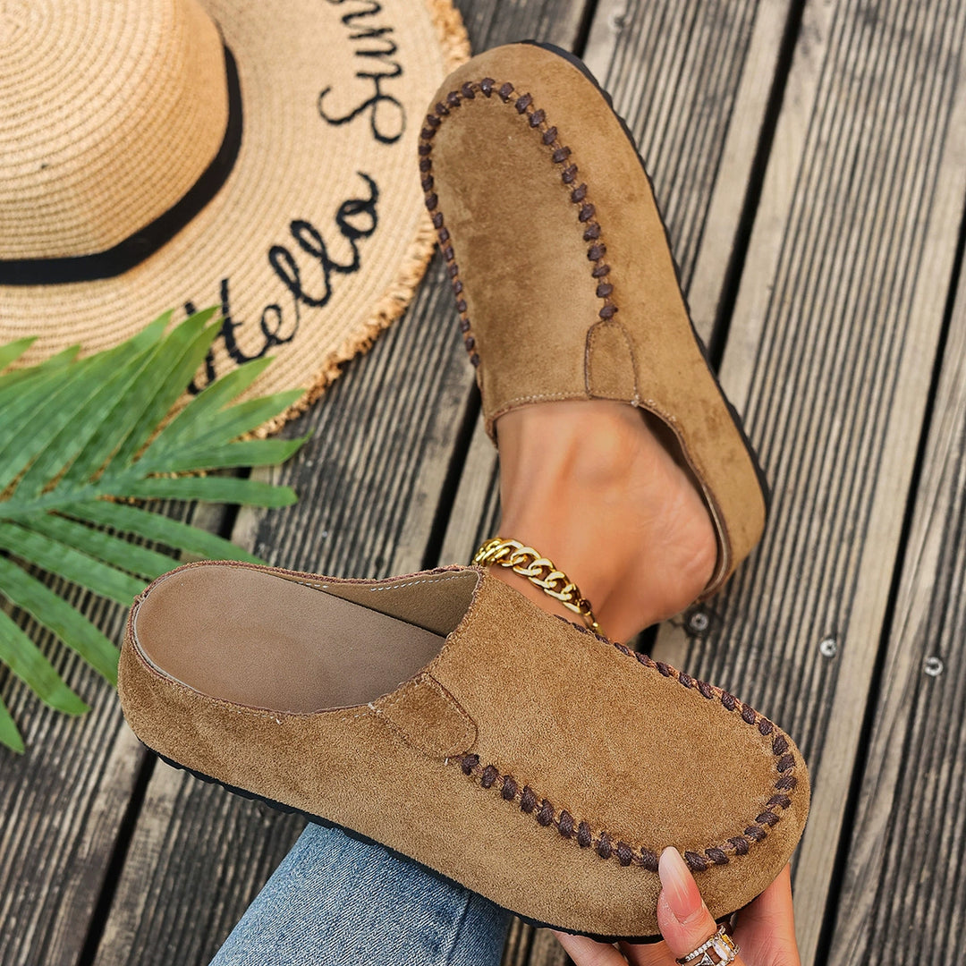 Tan mules held in hand next to a straw hat