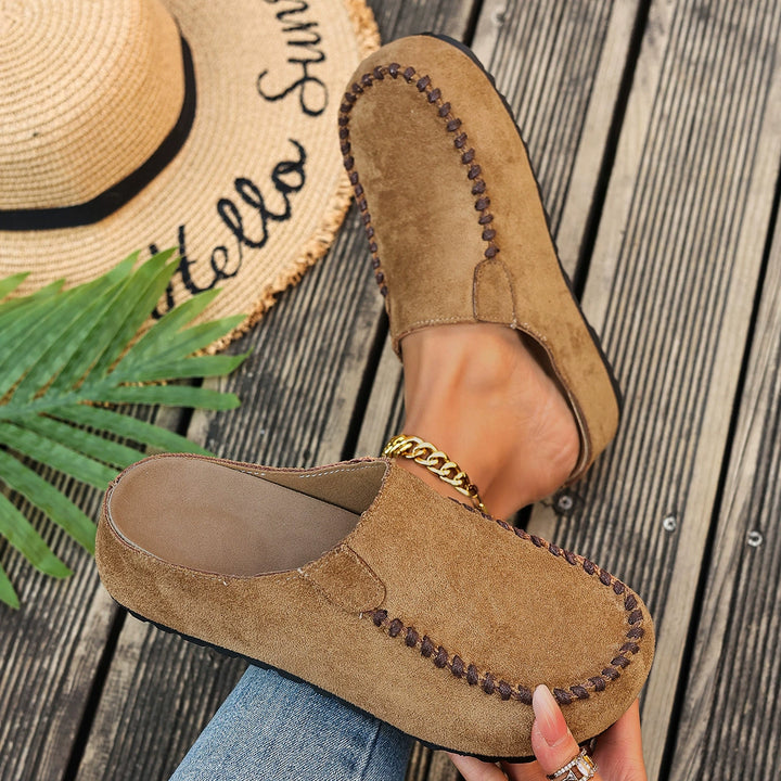 Tan mules held in hand next to a straw hat