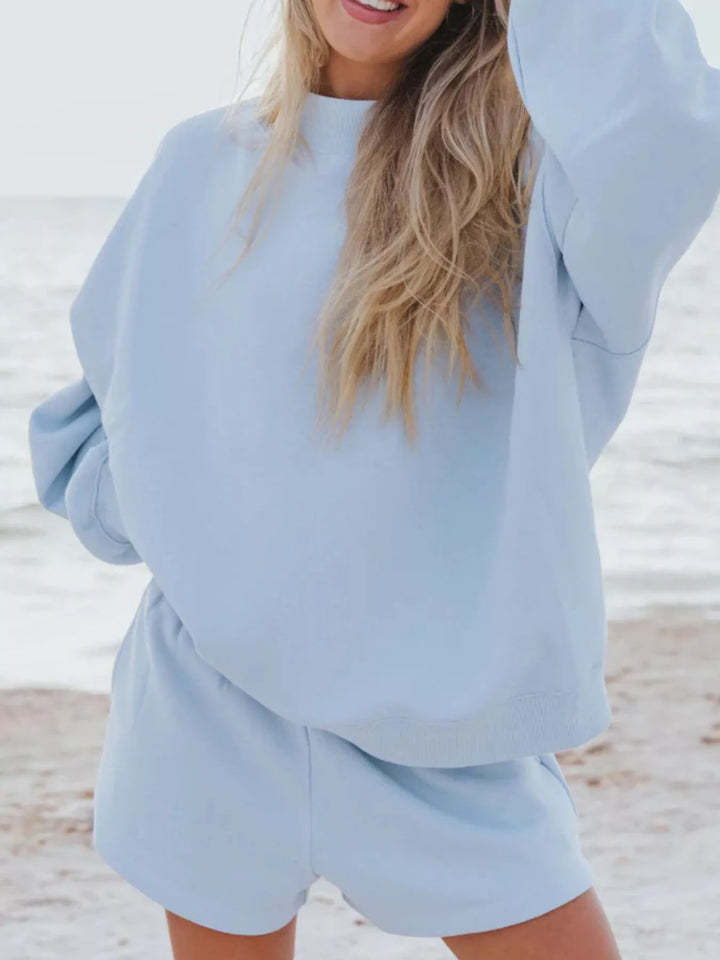 Sky blue oversized sweatshirt and shorts set, one arm raised, beach backdrop