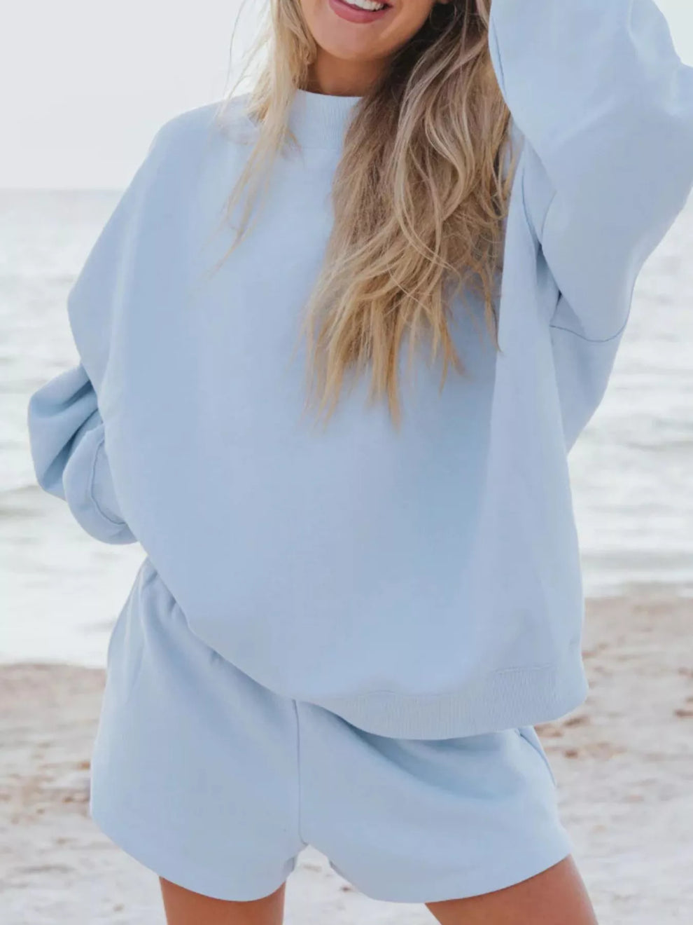 Sky blue oversized sweatshirt and shorts set, one arm raised, beach backdrop
