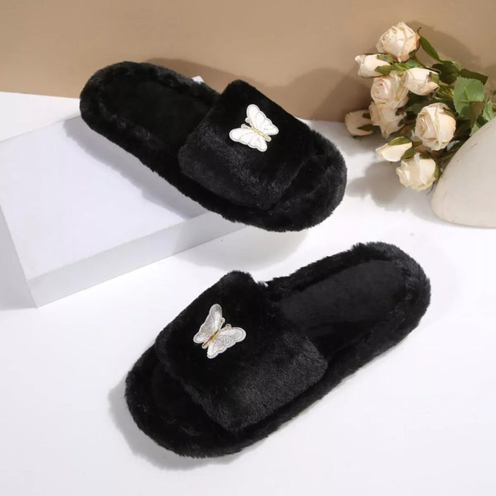 Two black plush butterfly slide slippers from above beside white flowers