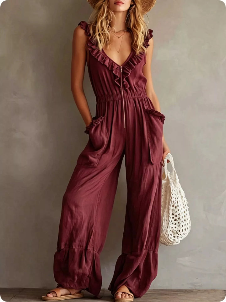 Burgundy wide leg jumpsuit with ruffle V neck, standing with a mesh bag