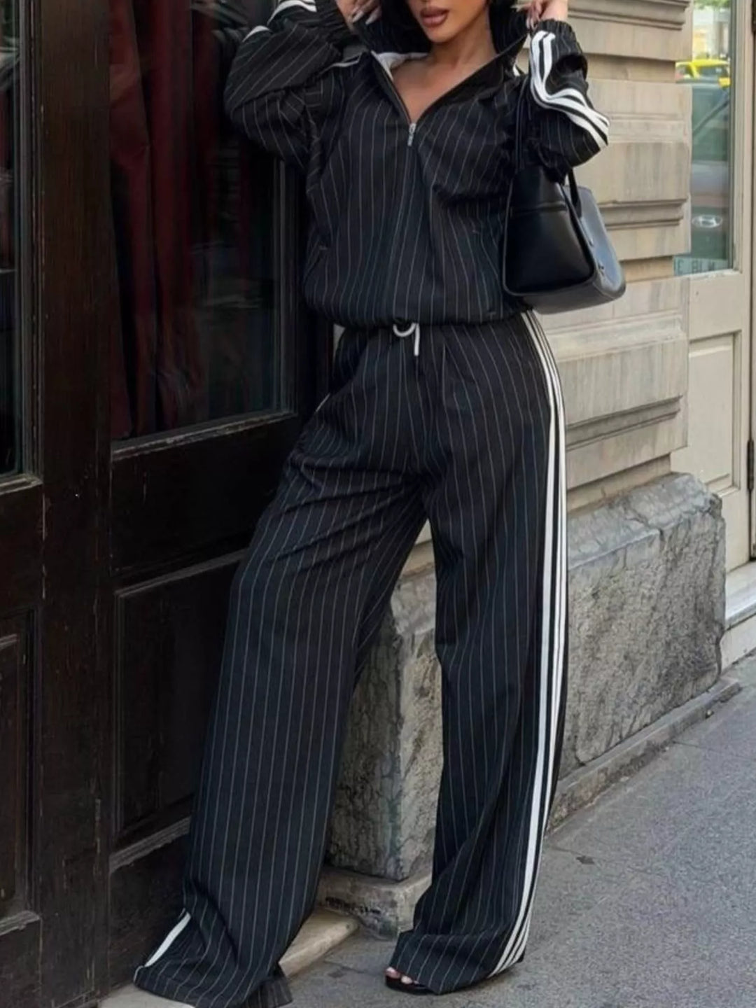 Black pinstripe zip jacket and wide leg pants, leaning on a doorway with a handbag